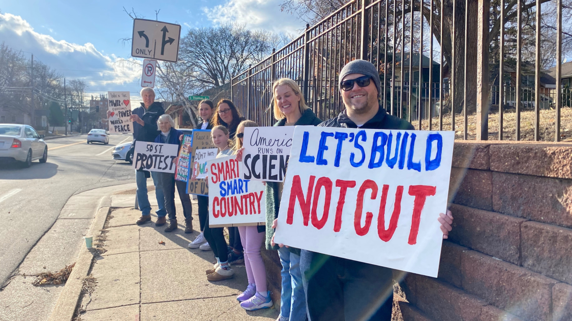 a photo of people standing on a sidewalk holding signs that read: Let's build not cut. A neighborhood demonstration/protest.