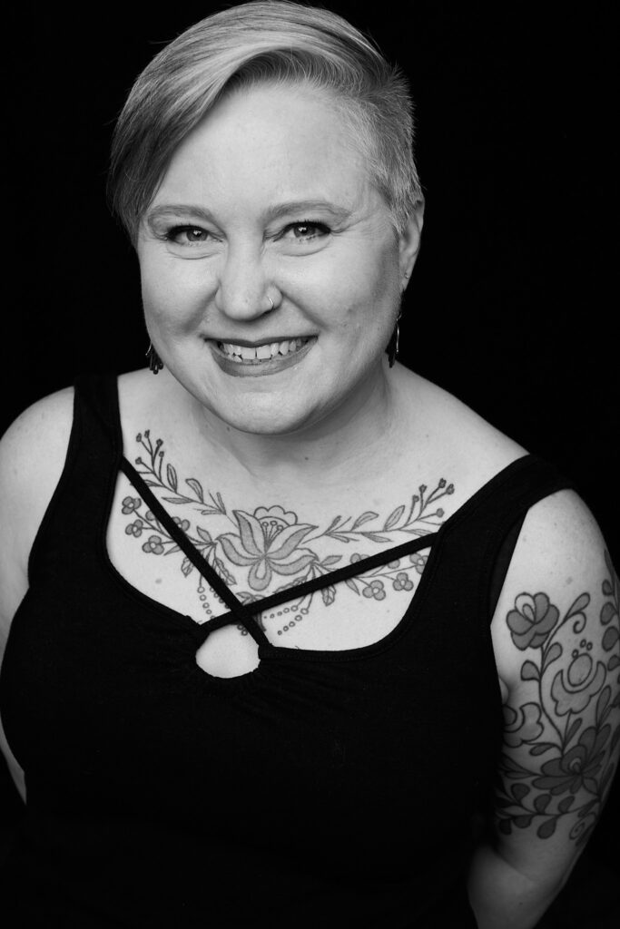 Headshot of Jeni Kolstad in black and white. She has short hair and is smiling at the camera wearing a black tank top with tattoos covering her chest and upper arm.