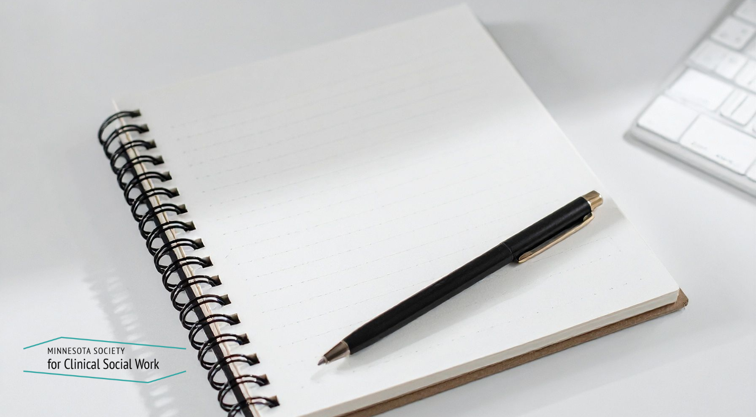 an image of a spiral bound notebook on a desk with white paper and a black pen laying on top