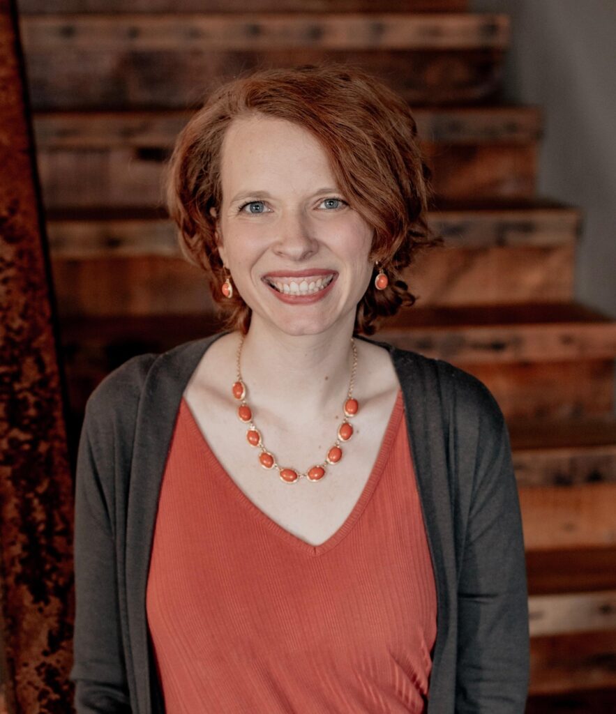 an image of Lindsey Peterson smiling at the camera with short red hair, orange necklace and an orange shirt